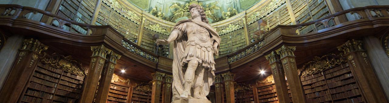  Prunksaal in der österreichischen Nationalbibliothek mit einer Statue in der Mitte des Raumes, im Hintergrund eine Balustrade überall sind Regale mit alten Büchern