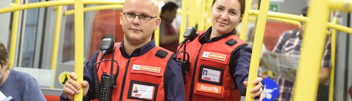 zwei Wiener Linien Mitarbeiter mit roten Sicherheitswesten stehen in einem U-Bahn-Zug und lächeln in die Kamera