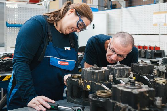 eine Frau in Arbeitskleidung und Schutzbrille mit Werkstücken in einer Werkstätte