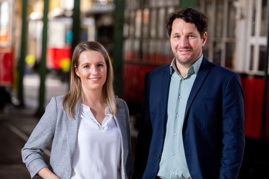 Zwei Personen in Businesskleidung (ein Mann und eine Frau) stehen vor einer Straßenbahn